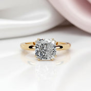 A yellow gold solitaire engagement ring featuring a cushion-cut diamond center stone, shown on a clean white background with a soft reflection below.