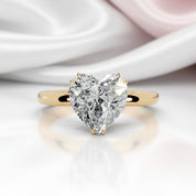 A heart-shaped diamond solitaire ring set on a yellow gold band, displayed on a reflective white surface with soft white and pale pink satin fabric in the background.