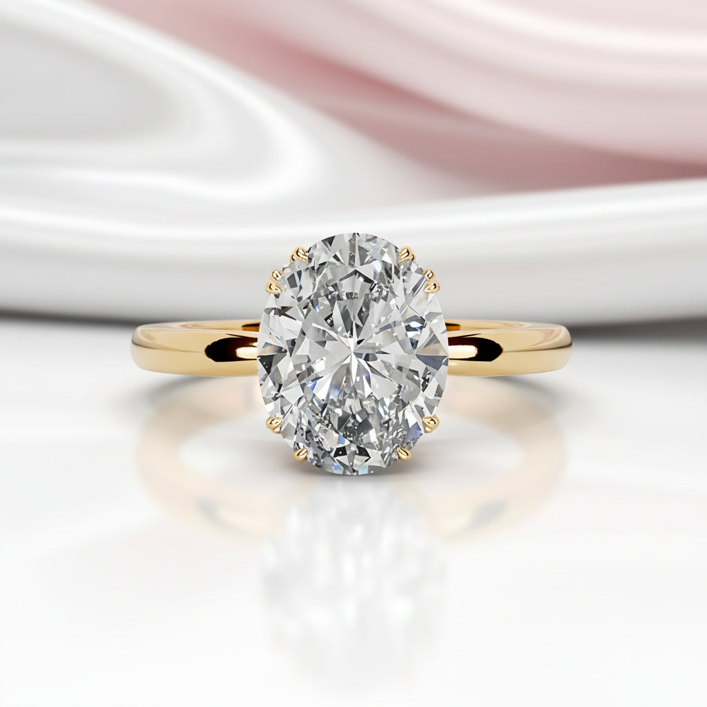 A yellow gold engagement ring featuring a large oval-cut diamond in a prong setting, resting on a reflective white surface with a soft pink satin fabric blurred in the background.