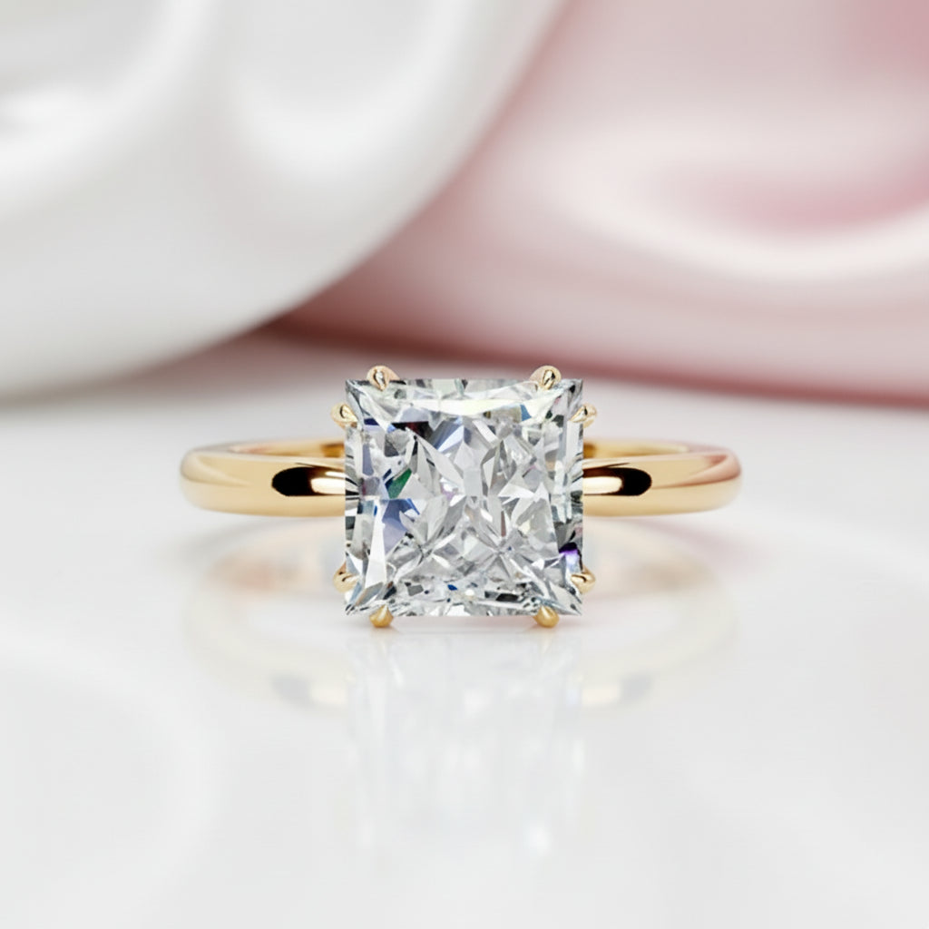 A radiant cut diamond engagement ring with a yellow gold band, shown in a close-up studio shot with a soft white and pink satin background.
