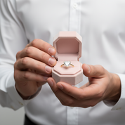 A close-up of a person in a white dress shirt holding an open light pink jewelry box that displays a gold ring with a heart-shaped clear gemstone.