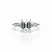 An emerald-cut diamond ring with baguette side stones in a silver-toned setting, centered on a reflective white surface.