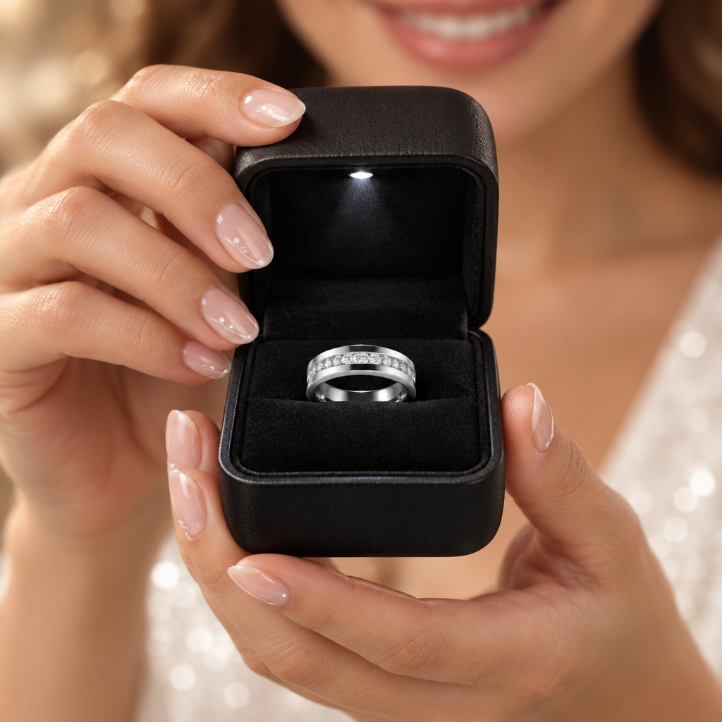 A close-up lifestyle shot of a woman holding an open black ring box with a silver band ring inside, set against a soft, blurred background of her face and white dress.
