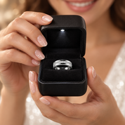 A close-up lifestyle shot of a woman holding an open black ring box with a silver band ring inside, set against a soft, blurred background of her face and white dress.