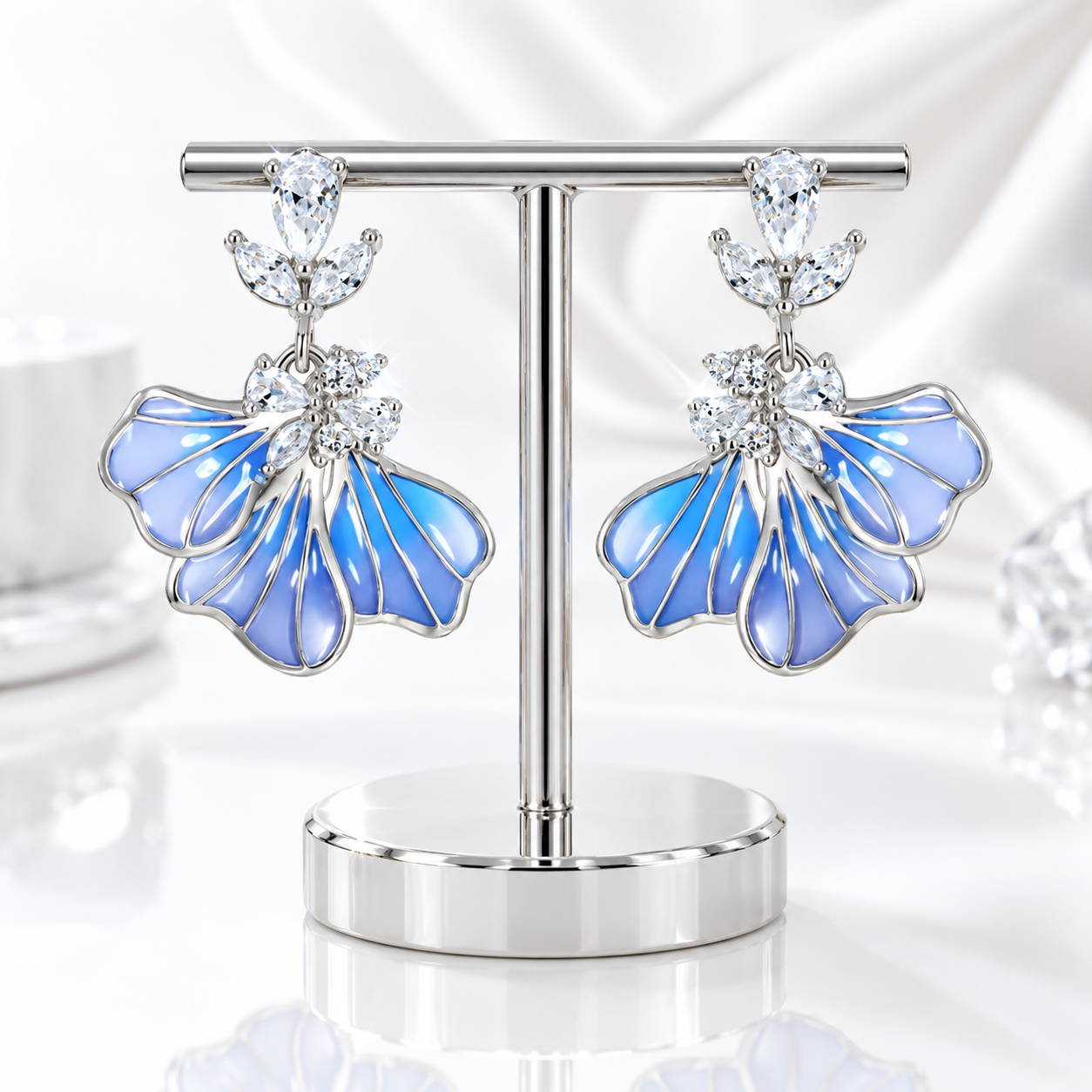 A pair of dangling statement earrings displayed on a silver T-stand. The earrings have iridescent light blue and lavender-blue petal-shaped enamel sections outlined in silver, with clear gemstone clusters at the top and center.