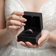 A close-up of a person holding an open black ring box with a ring inside. The person is wearing a sparkling silver dress, and the ring appears to be a silver band with a red and black center section.
