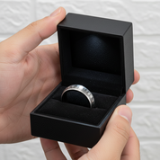 Hand holding a black jewelry box with a silver ring inside against a neutral background