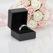 Black jewelry box with a ring inside, placed on a white surface with pink and white flowers in the background.