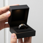 Hand holding a black jewelry box with a ring inside on a light gray background