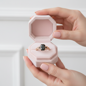 A hand holds an open pale pink jewelry box containing a ring with a dark rectangular stone and a rose-gold-toned band with small clear stones.