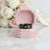 A black carbon-fiber-patterned ring with a small clear gemstone, displayed inside an open pink octagonal ring box on a white marble surface with blurred pink roses in the background.