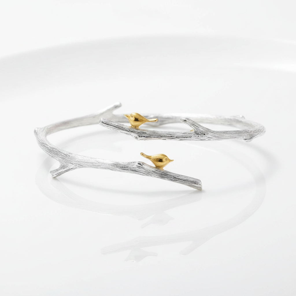 A silver-toned open cuff bracelet designed to look like a textured tree branch, featuring two small gold-toned birds perched on it, photographed on a clean white reflective surface.