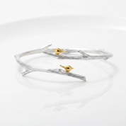 A silver-toned open cuff bracelet designed to look like a textured tree branch, featuring two small gold-toned birds perched on it, photographed on a clean white reflective surface.