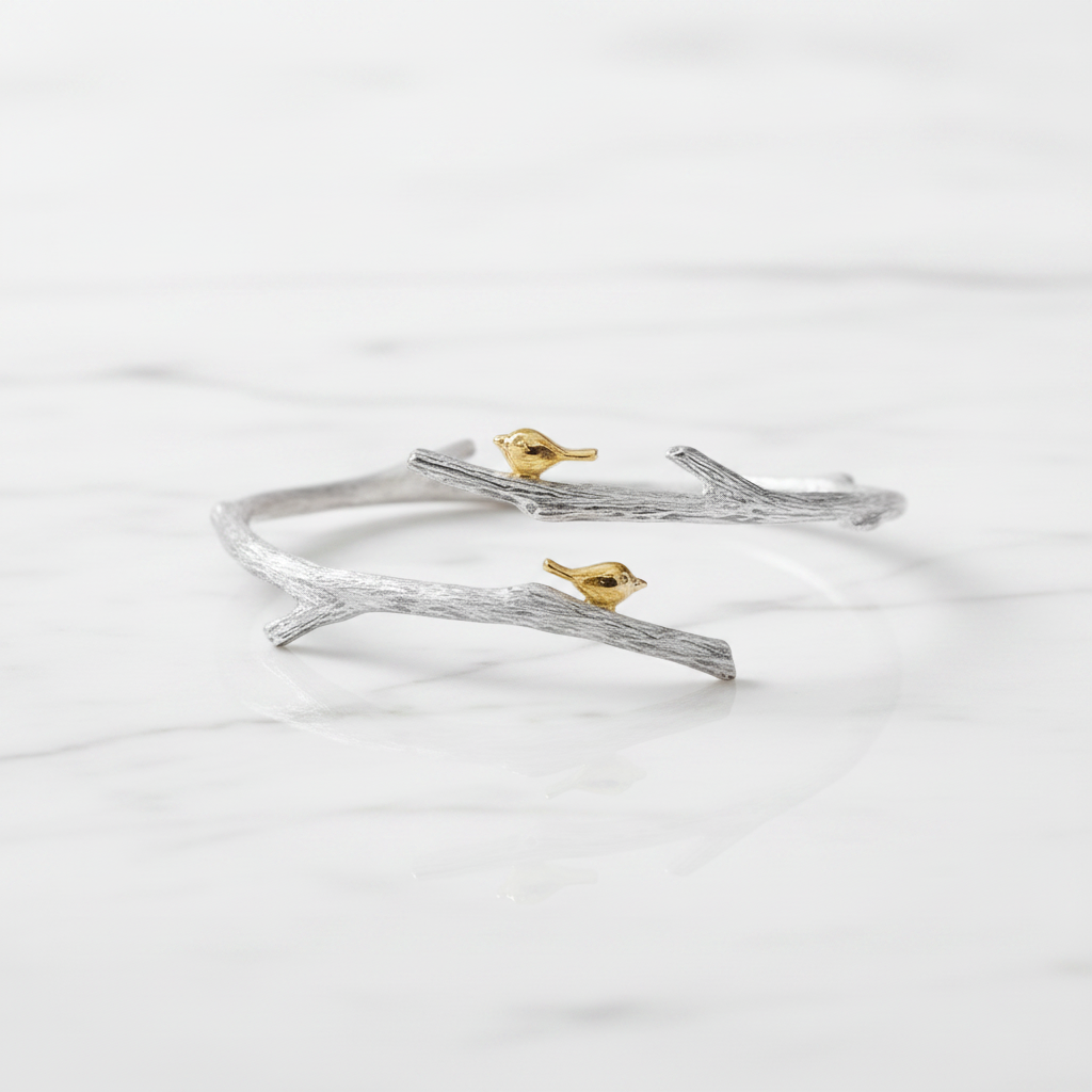 A silver branch-shaped cuff bracelet with two small gold birds perched on it, displayed on a white marble surface.