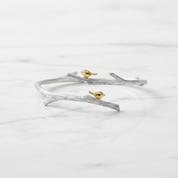 A silver branch-shaped cuff bracelet with two small gold birds perched on it, displayed on a white marble surface.