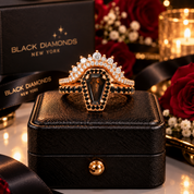 A coffin-shaped ring with a dark smoky gemstone and black gemstone accents, set in a rose gold-toned setting with clear stones, displayed on a black textured jewelry box. The background features a black 'BLACK DIAMONDS NEW YORK' box, red roses, and warm candlelight.