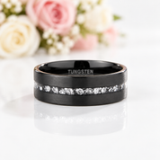 A black tungsten ring band with a channel-set row of clear stones, shown on a white reflective surface with soft pink and white flowers blurred in the background. The inside of the band is engraved with the word 'TUNGSTEN'.