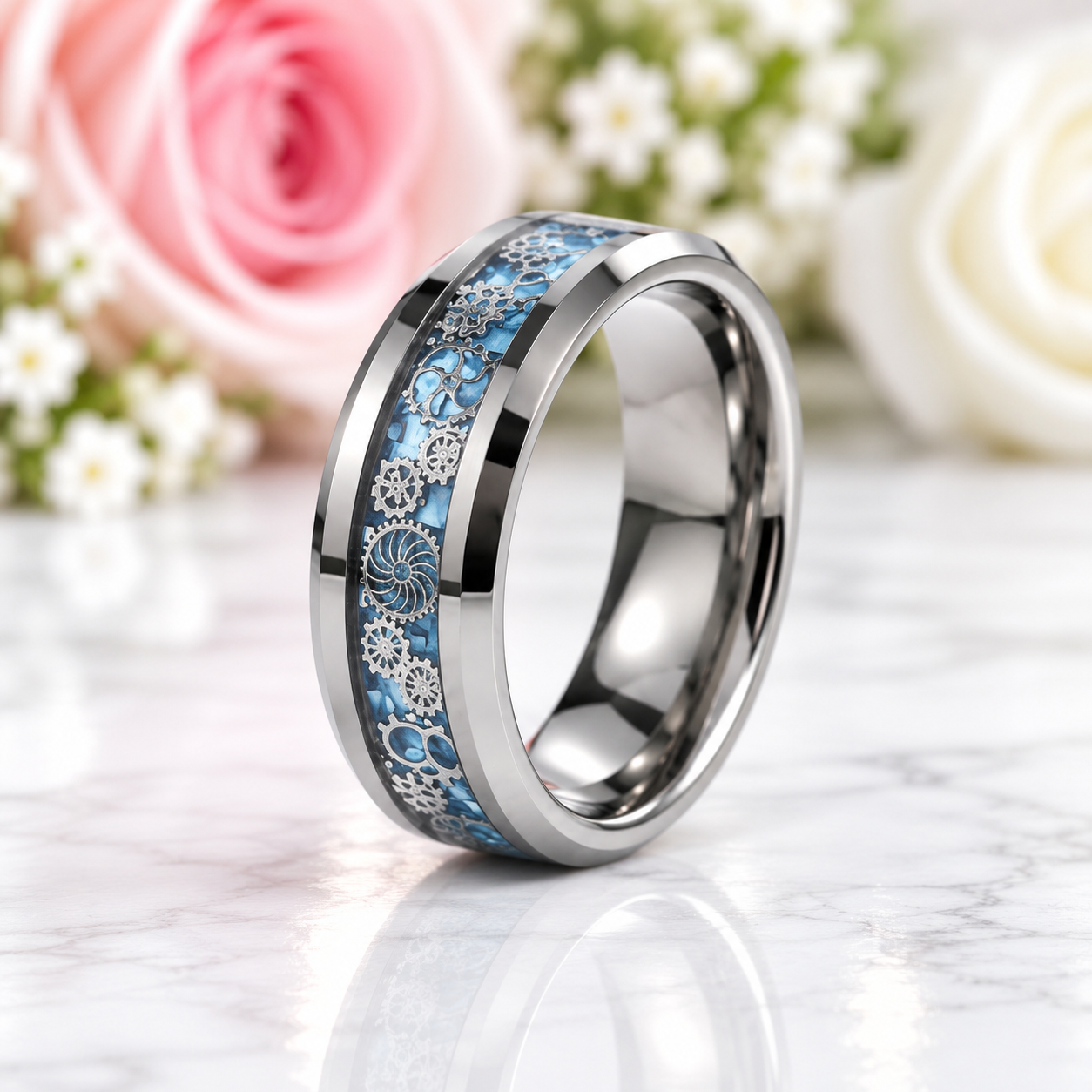 A polished silver-toned band ring with a decorative inlay of light blue and silver floral/leaf patterns, displayed on a white marble surface with blurred pink and white roses in the background.