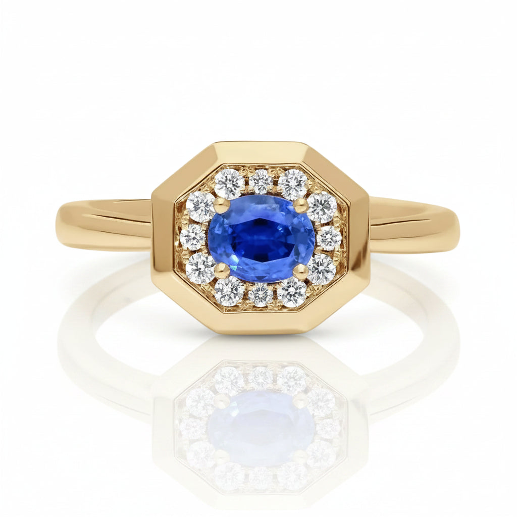 An octagonal yellow gold ring featuring a central oval blue sapphire gemstone surrounded by a halo of small round clear diamonds, set against a clean white background with a soft reflection.
