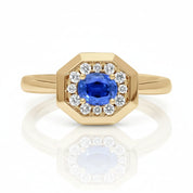 An octagonal yellow gold ring featuring a central oval blue sapphire gemstone surrounded by a halo of small round clear diamonds, set against a clean white background with a soft reflection.