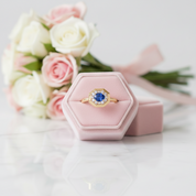 A gold ring with a blue and white gemstone centerpiece is displayed in a pink hexagonal ring box, with a blurred bouquet of pink and white roses in the background.