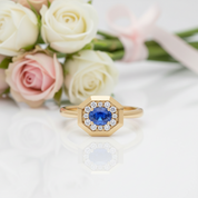 A gold hexagonal ring with a central blue sapphire and a halo of small clear stones, styled on a white reflective surface with soft pink and white roses in the background.
