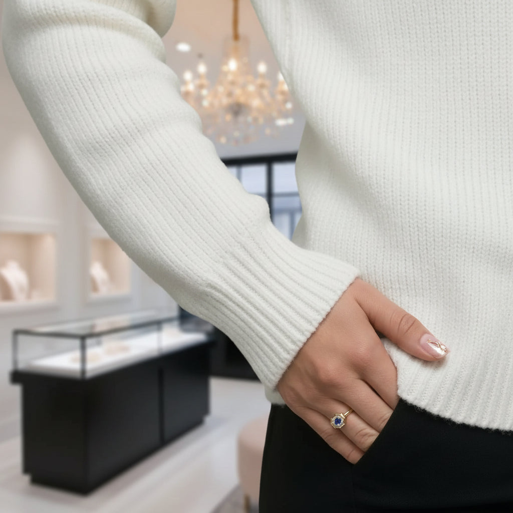 A close-up of a person wearing a ribbed white knit sweater and a black skirt, with one hand in the pocket and wearing a gold ring with a blue and clear gemstone. The background shows a softly blurred boutique interior with display cases and a chandelier.