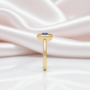 A gold ring with a small round blue gemstone, standing upright against a soft, draped white and pale pink satin fabric background.