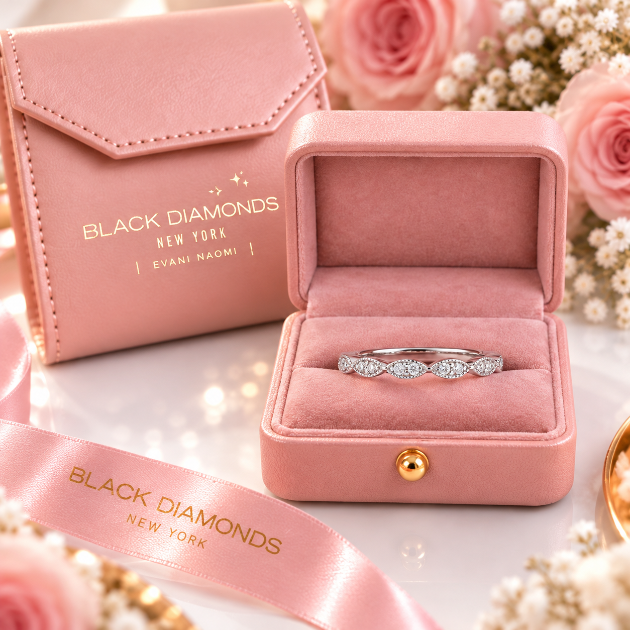 A silver band ring with five round-cut clear stones displayed in an open pink velvet jewelry box. A matching pink envelope-style box and pink satin ribbon with gold 'BLACK DIAMONDS NEW YORK' branding are arranged around it on a light surface with soft pink roses in the background.
