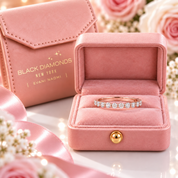 A rose gold band ring with a row of small clear stones sits in an open pink velvet ring box. A branded pink jewelry box and satin ribbon are visible in the background with soft floral decor.
