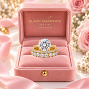 A yellow gold engagement ring with a large round-cut diamond and a matching diamond-studded band, presented in an open pink velvet Black Diamonds New York box with pink roses and ribbon around it.