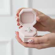 Hand holding a pink jewelry box with a diamond ring inside against a light background