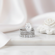 A silver-toned engagement ring with a large oval-cut clear center stone, stacked with matching diamond-studded bands, displayed on a white marble surface with a soft white rose prop in the background.