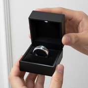 Person holding a black jewelry box with a silver ring inside against a neutral background