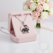 A silver-toned pendant necklace with a detailed fairy illustration on a crescent moon, displayed in an open light pink jewelry box. A bouquet of pink and white roses is softly blurred in the background.