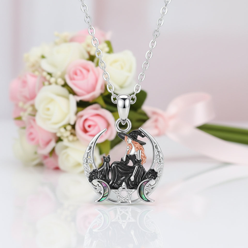 A silver pendant necklace featuring a witch sitting on a crescent moon with a black cat, displayed on a reflective white surface with a blurred bouquet of pink and white roses in the background.