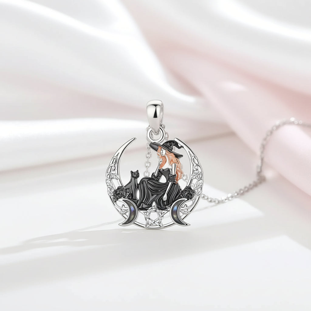 A silver-toned pendant necklace featuring a witch sitting on a crescent moon with black cats, accented with gold and iridescent blue details, displayed on draped white and pale pink satin fabric.