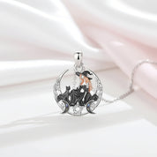 A silver-toned pendant necklace featuring a witch sitting on a crescent moon with black cats, accented with gold and iridescent blue details, displayed on draped white and pale pink satin fabric.