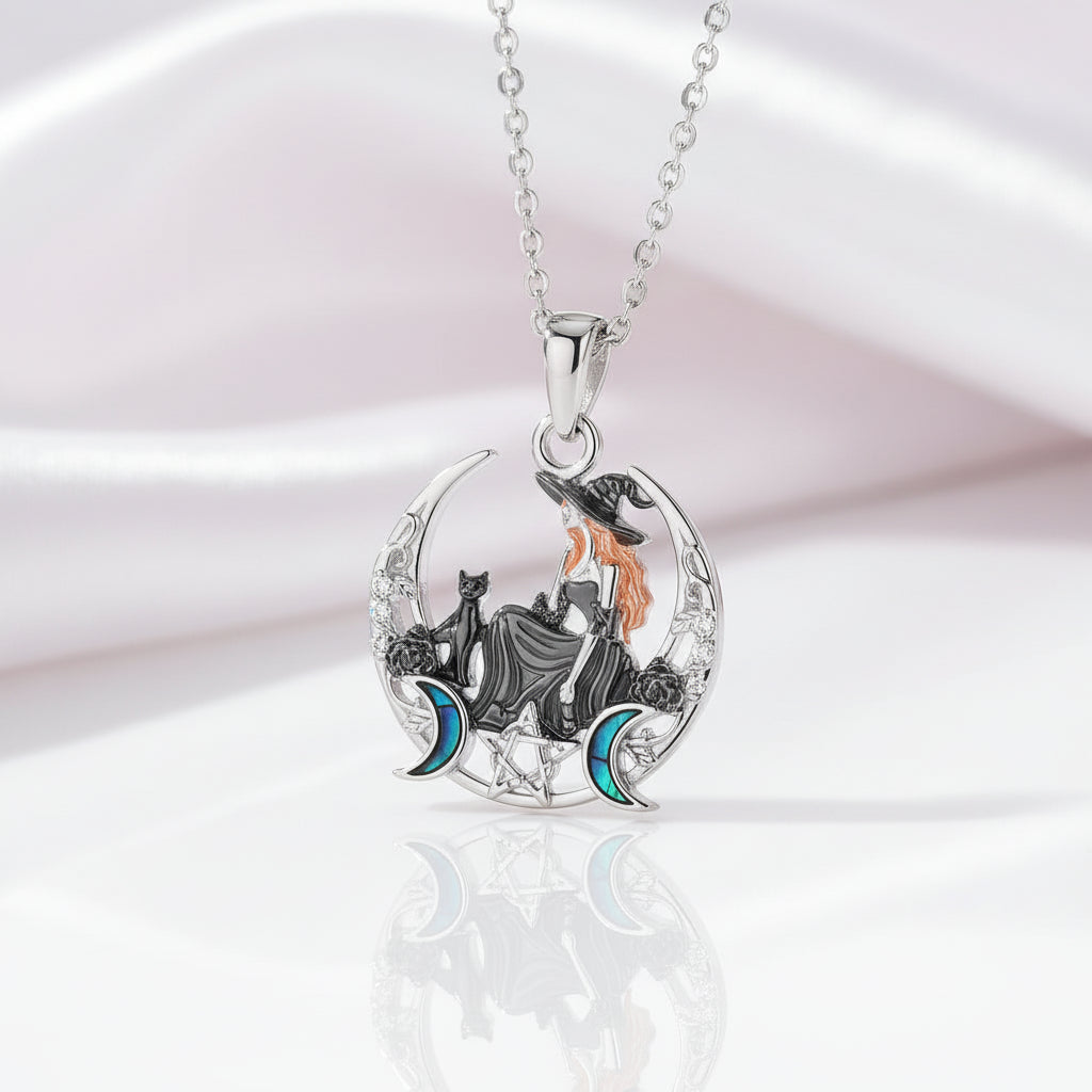 A silver pendant necklace featuring a witch sitting on a crescent moon with a black cat, accented with orange hair, blue moons, and clear crystals, displayed on a soft white satin background.