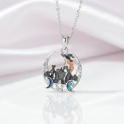 A silver pendant necklace featuring a witch sitting on a crescent moon with a black cat, accented with orange hair, blue moons, and clear crystals, displayed on a soft white satin background.