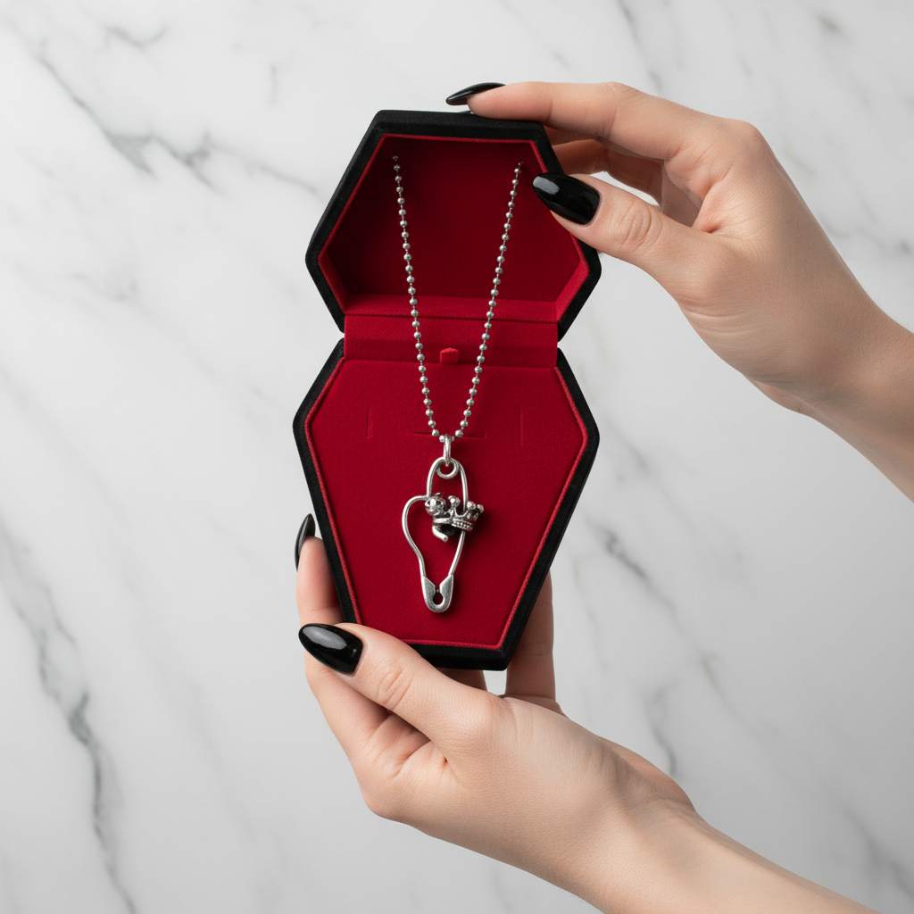 A silver ball-chain necklace with a safety-pin heart pendant and small skull charms, displayed inside an open black coffin-shaped box with a red velvet lining, held by two hands with black nail polish against a white marble background.