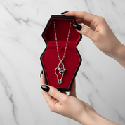 A silver ball-chain necklace with a safety-pin heart pendant and small skull charms, displayed inside an open black coffin-shaped box with a red velvet lining, held by two hands with black nail polish against a white marble background.