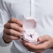 A person in a white button-up shirt holds an open light pink jewelry box displaying a silver-toned winged pendant with a purple and iridescent gemstone.