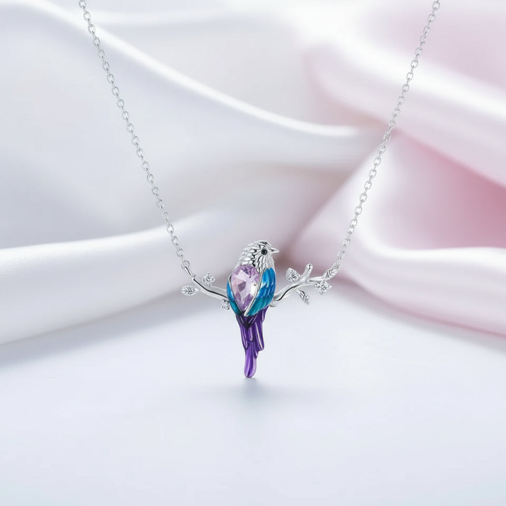 A silver necklace with a bird-shaped pendant featuring purple, blue, and pink crystal details, displayed on a soft white and pink satin background.