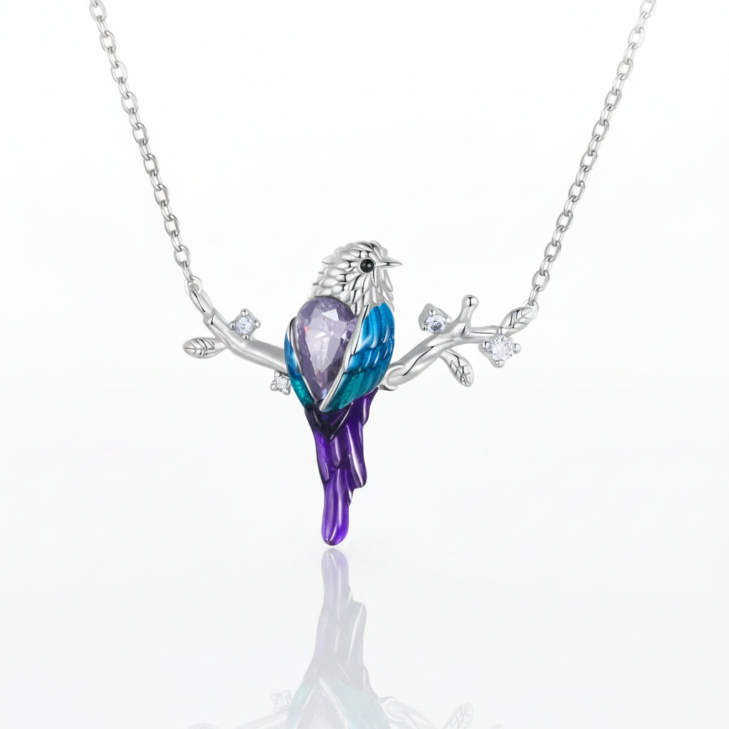 A silver-tone necklace featuring a bird pendant with a faceted purple crystal body, blue and purple enamel wings, and small clear stones on the branch, shown on a white background with a soft reflection.