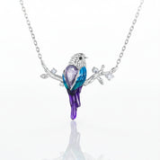 A silver-tone necklace featuring a bird pendant with a faceted purple crystal body, blue and purple enamel wings, and small clear stones on the branch, shown on a white background with a soft reflection.