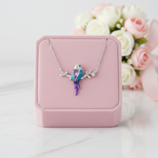 A silver necklace with a bird pendant featuring blue and purple crystal details, displayed on a pink jewelry box with blurred white and pink flowers in the background.