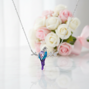 A silver necklace with a bird pendant made of iridescent blue and purple crystals, displayed against a soft-focus background of white and pink roses.