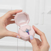 A silver necklace with a colorful bird pendant is displayed in an open pink jewelry box, held by two hands against a soft white background.