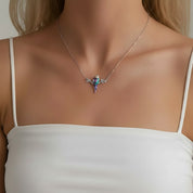 A close-up of a model wearing a delicate silver necklace with a small, iridescent butterfly pendant. The model is wearing a white spaghetti-strap top against a neutral light gray background.